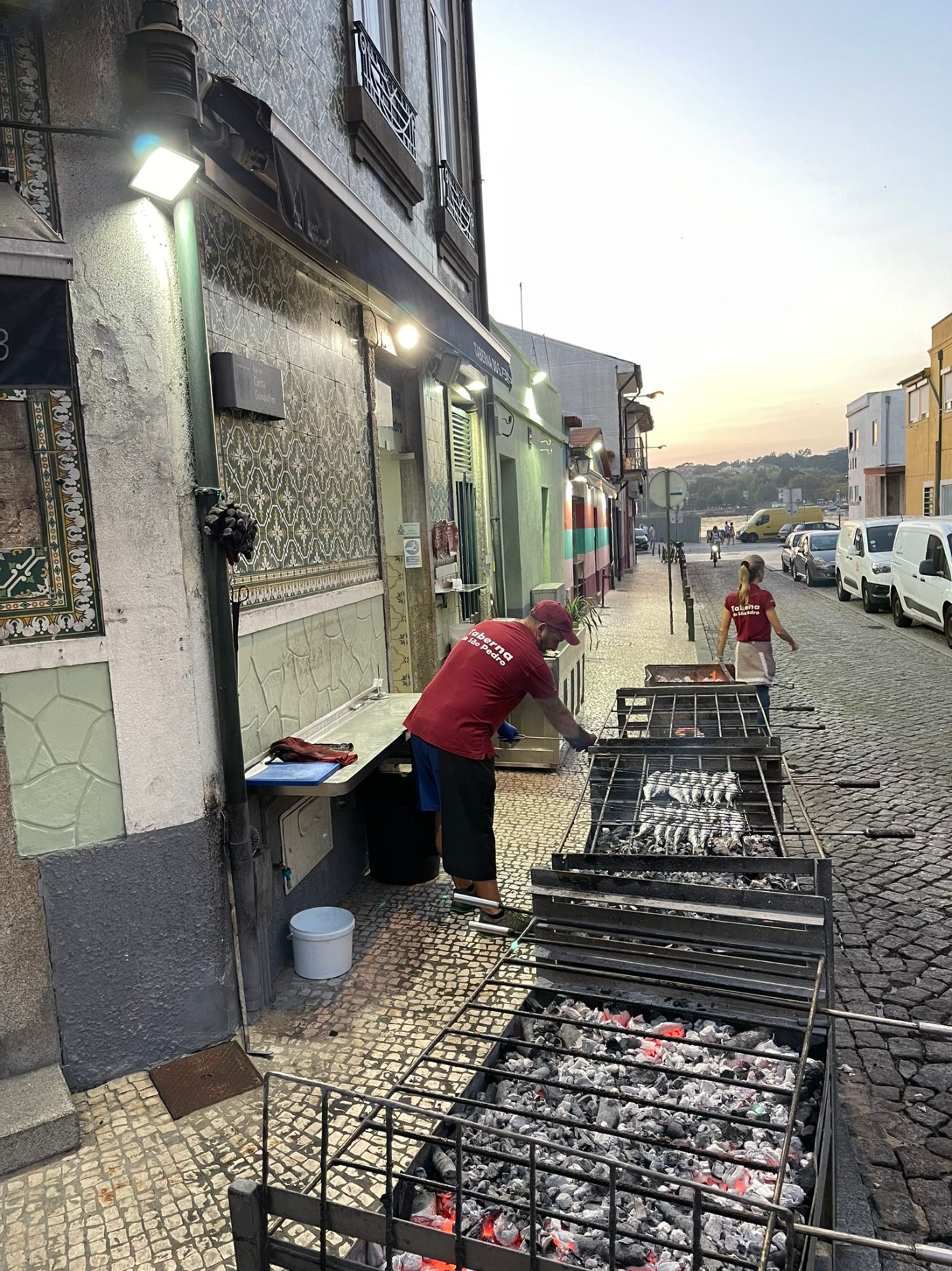 A row of charcoal grills set up on the cobbled street outside a Porto taberna, fish and sardines cooking over the coals